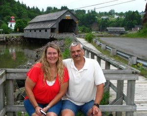 Bill Caswell and a friend sitting on a deck next to a boarded path, which leads to a covered bridge.